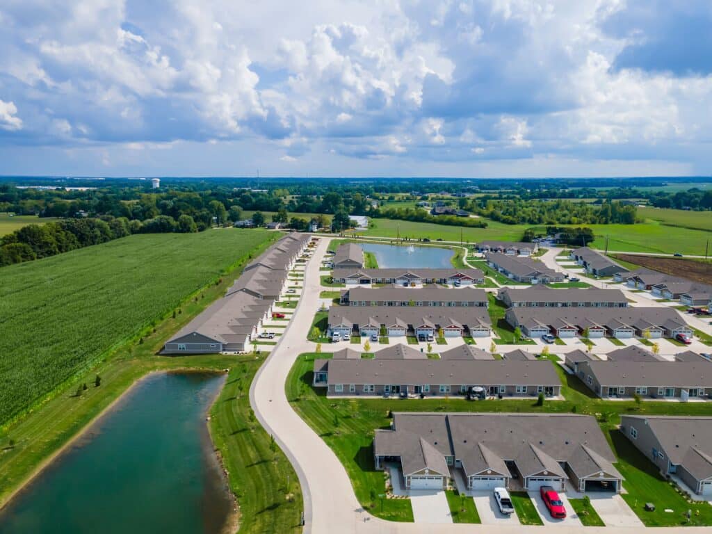 Aerial view of a suburban neighborhood with rows of houses, roads, ponds, and surrounding green fields under a partly cloudy sky.