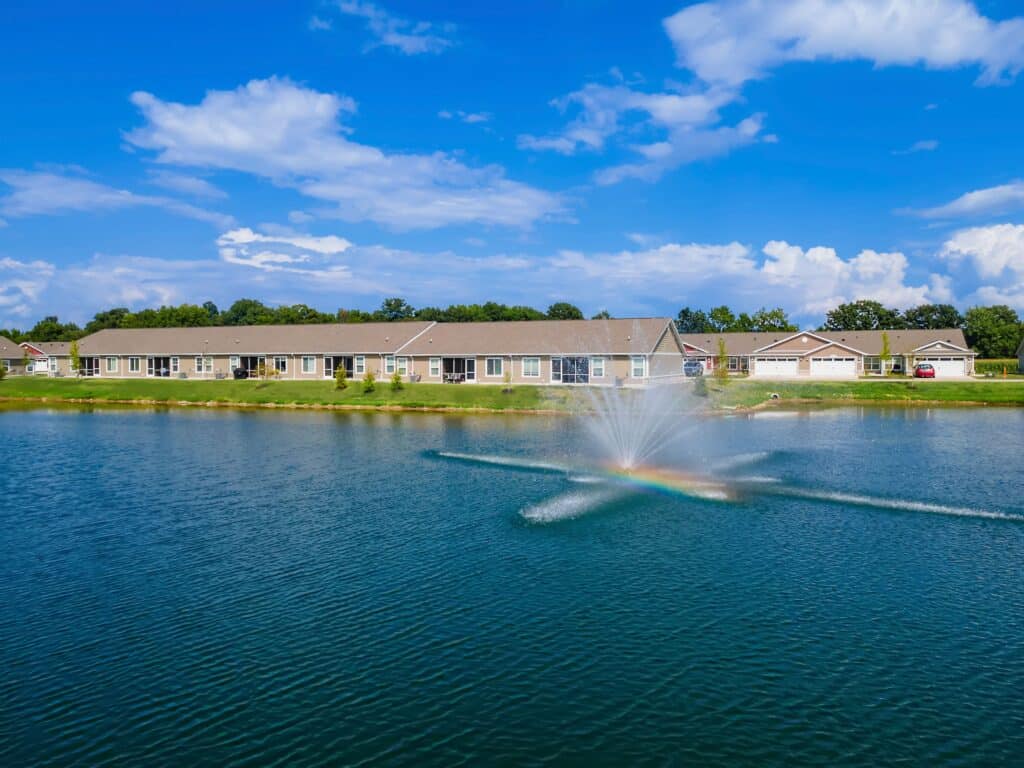 A row of single-story houses faces a pond with a central water fountain. A faint rainbow is visible in the fountain spray under a bright blue sky with scattered clouds.