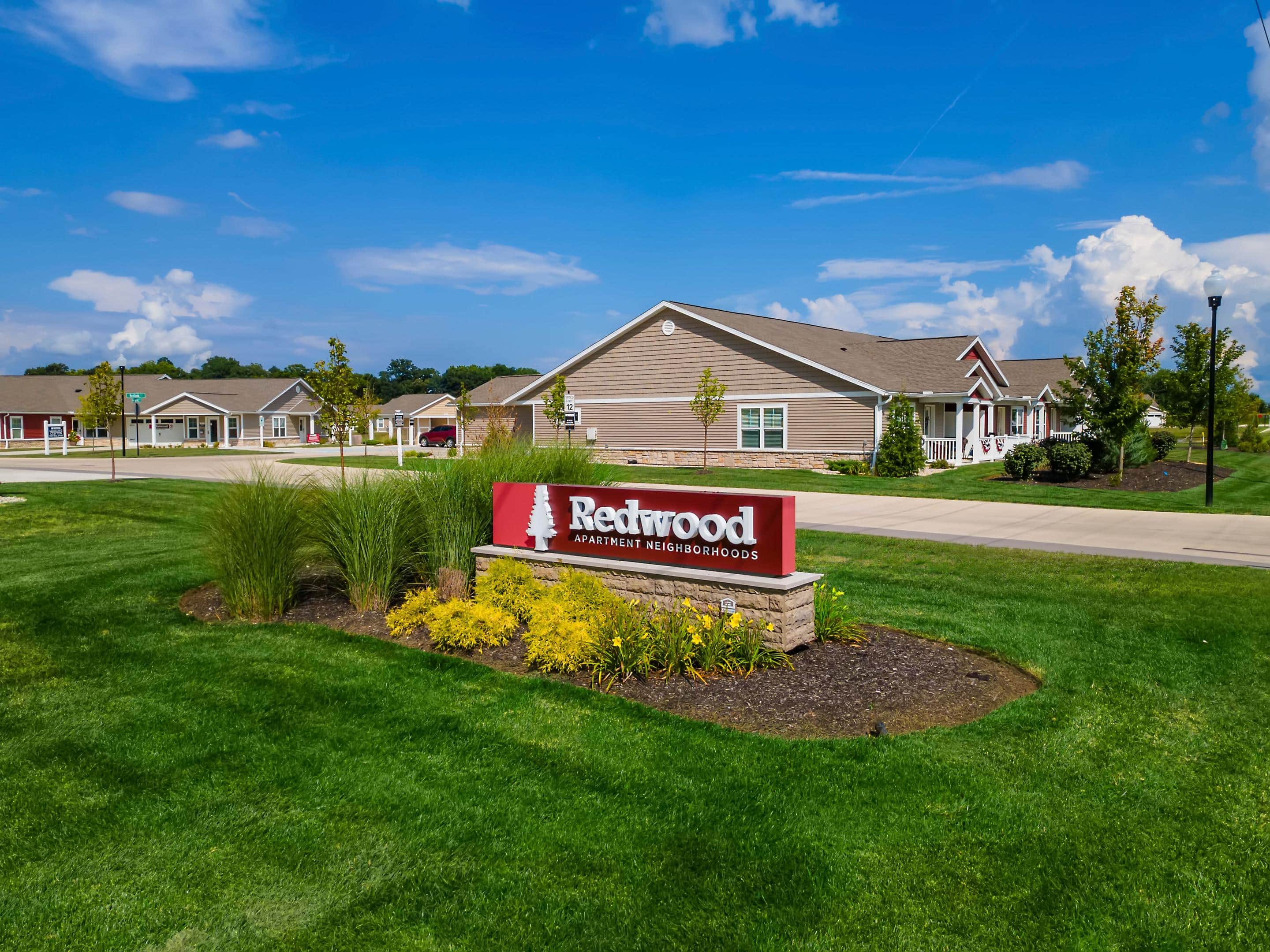 A landscaped sign reading "Redwood Apartment Neighborhoods" stands on a green lawn in front of single-story residential buildings under a blue sky.