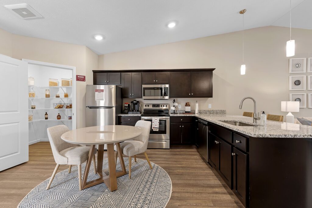 Modern kitchen with dark cabinets, stainless steel appliances, round dining table with two chairs, granite countertops, and a visible walk-in pantry.