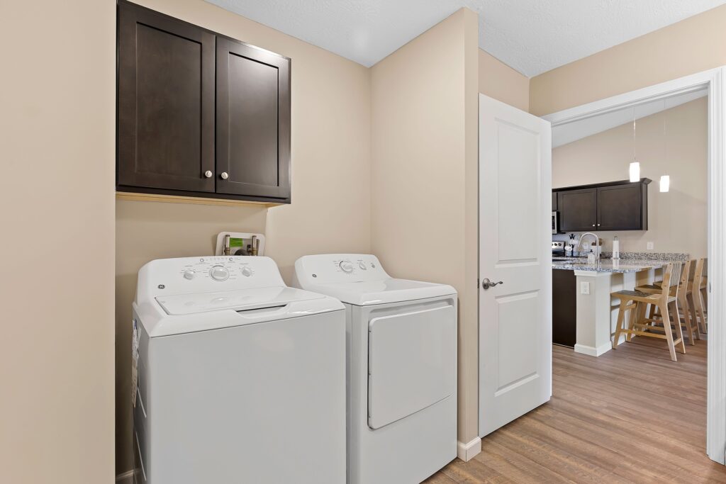 Laundry room with a washing machine and dryer side by side, a cabinet above, beige walls, and a view into a kitchen with barstools and pendant lights.