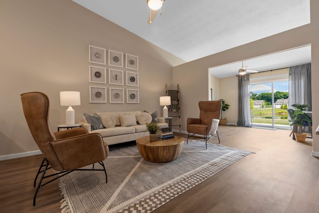 Modern living room with neutral decor, brown armchairs, a beige sofa, round wooden coffee table, floor lamp, and large window with outdoor view. Framed art displayed on the wall.