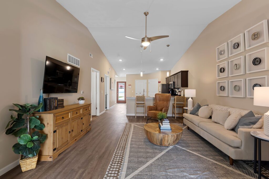 Modern living room and kitchen with wood floors, beige walls, a sofa, TV, dining area, and wall art; ceiling fan and indoor plant in view.