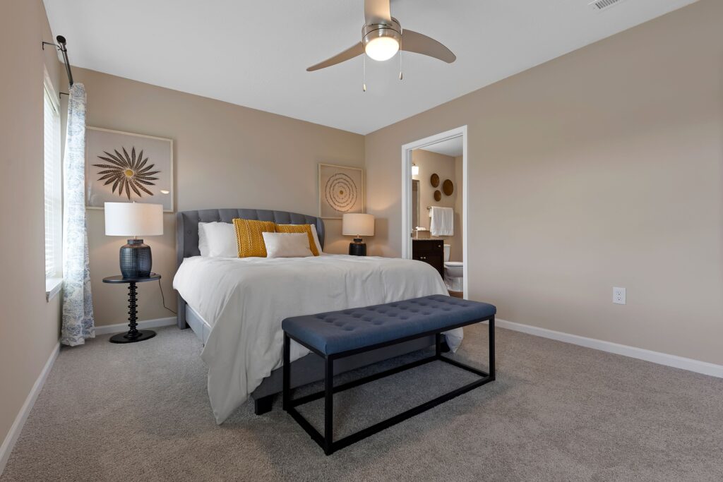 A neatly arranged bedroom with a bed, blue bench, two lamps, wall art, and an open doorway leading to a bathroom. Neutral walls and carpeted floor.