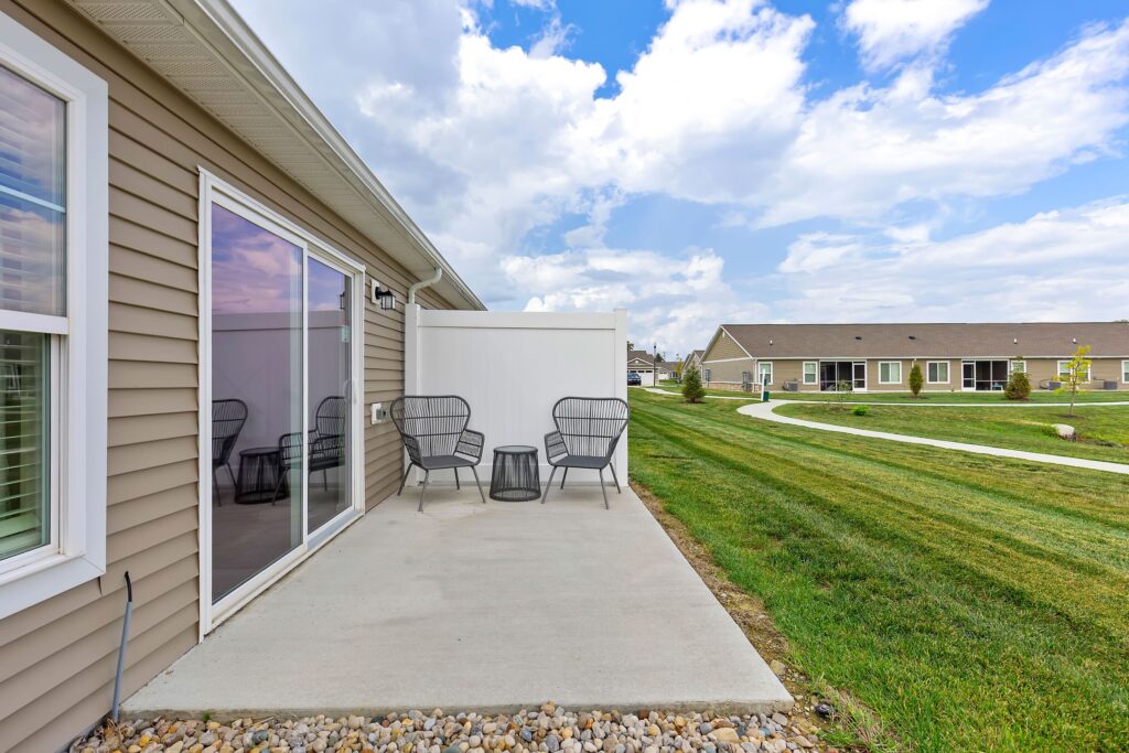 A concrete patio with three black chairs and a small table sits next to a tan house, overlooking a grassy yard with a sidewalk and other homes in the background under a partly cloudy sky.