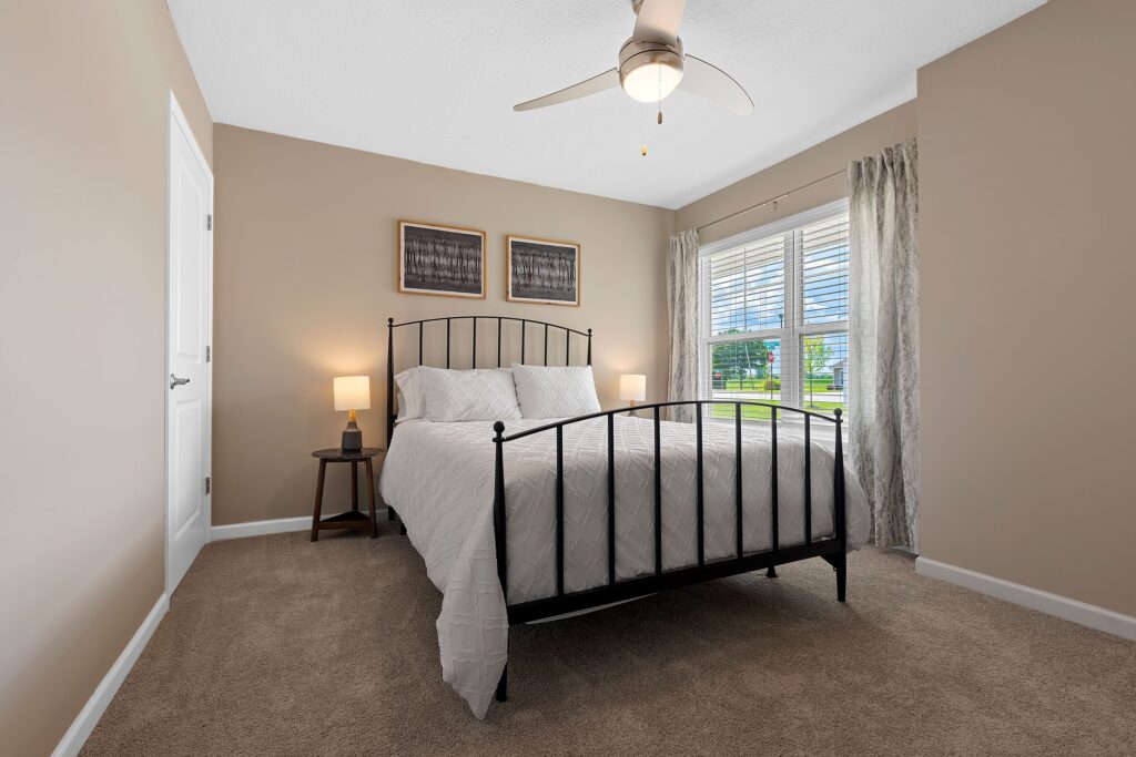 A bedroom with a black metal bed frame, white bedding, two bedside tables with lamps, beige walls, carpeted floor, and a window with curtains letting in natural light.