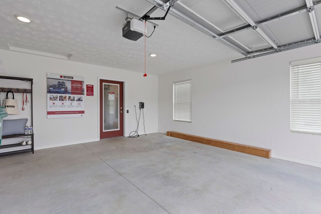 A clean, empty garage with white walls, two windows, a red door, a wall-mounted shelf, an overhead garage door opener, and a large poster on the wall.