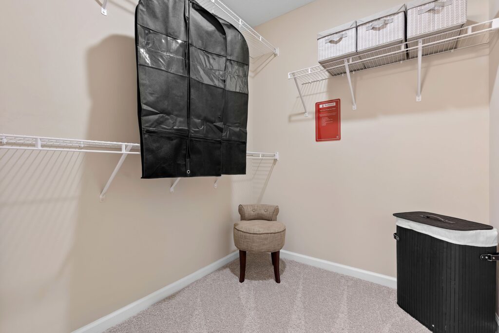 A walk-in closet with beige walls, white wire shelves, a black garment bag, two storage baskets, a beige upholstered stool, and a black laundry hamper.
