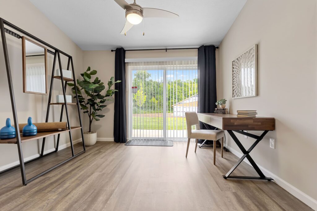Modern home office with wood floors, a desk and chair near sliding glass doors, a tall plant, metal shelving, and wall art, with natural light coming in from outside.