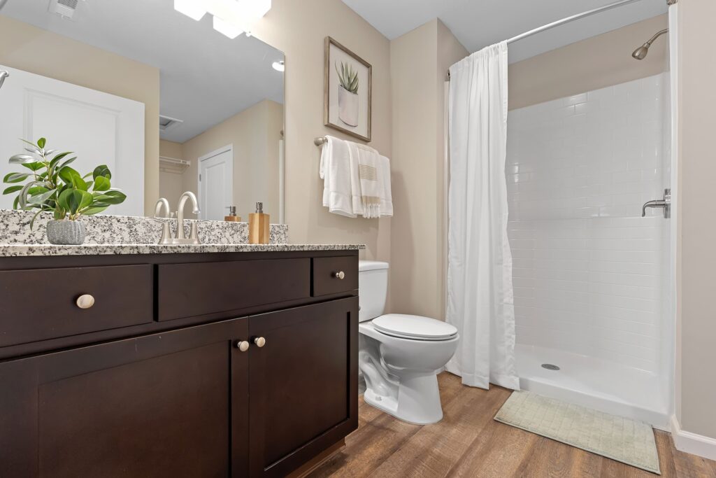 Modern bathroom with a granite countertop, dark wood vanity, white toilet, walk-in shower with white curtain, towels, potted plant, and framed wall art.