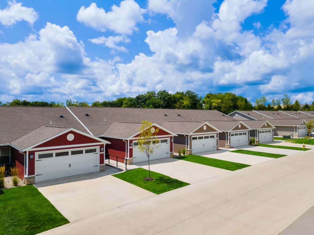 Row of single-story houses with attached garages, neatly trimmed lawns, and a wide driveway under a partly cloudy sky.