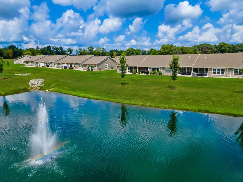 A row of single-story houses faces a grassy lawn and a pond with a water fountain, under a partly cloudy sky.
