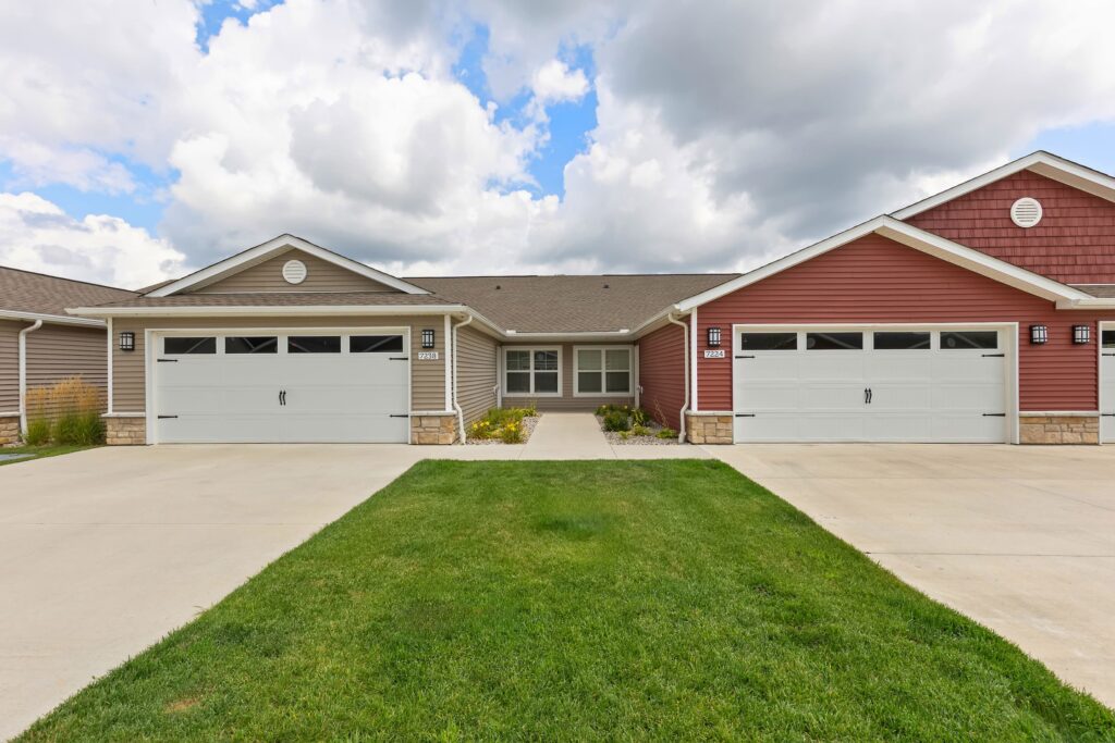 A duplex building with two garages, beige and red siding, stone accents, a shared walkway, and a green lawn under a cloudy sky.