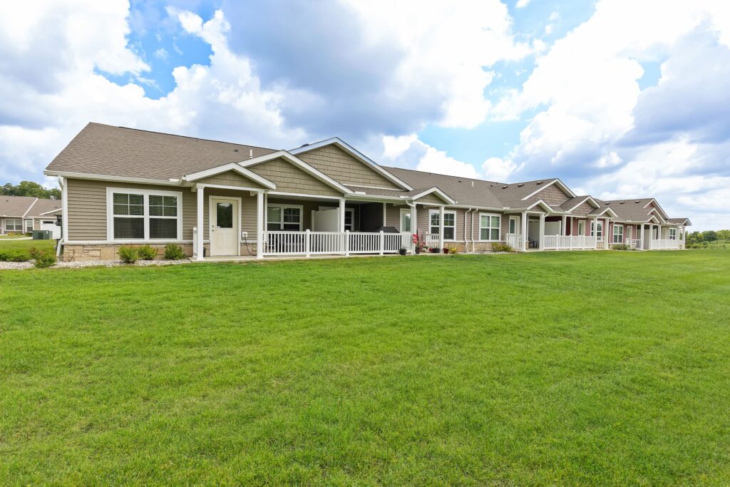 Single-story row of suburban homes with front porches, beige siding, and a large grassy lawn under a partly cloudy sky.