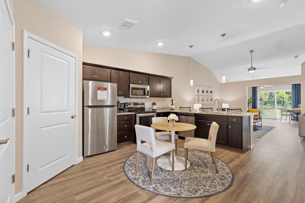 Open kitchen and dining area with stainless steel appliances, dark cabinets, a round table with four chairs, and a living room visible in the background.
