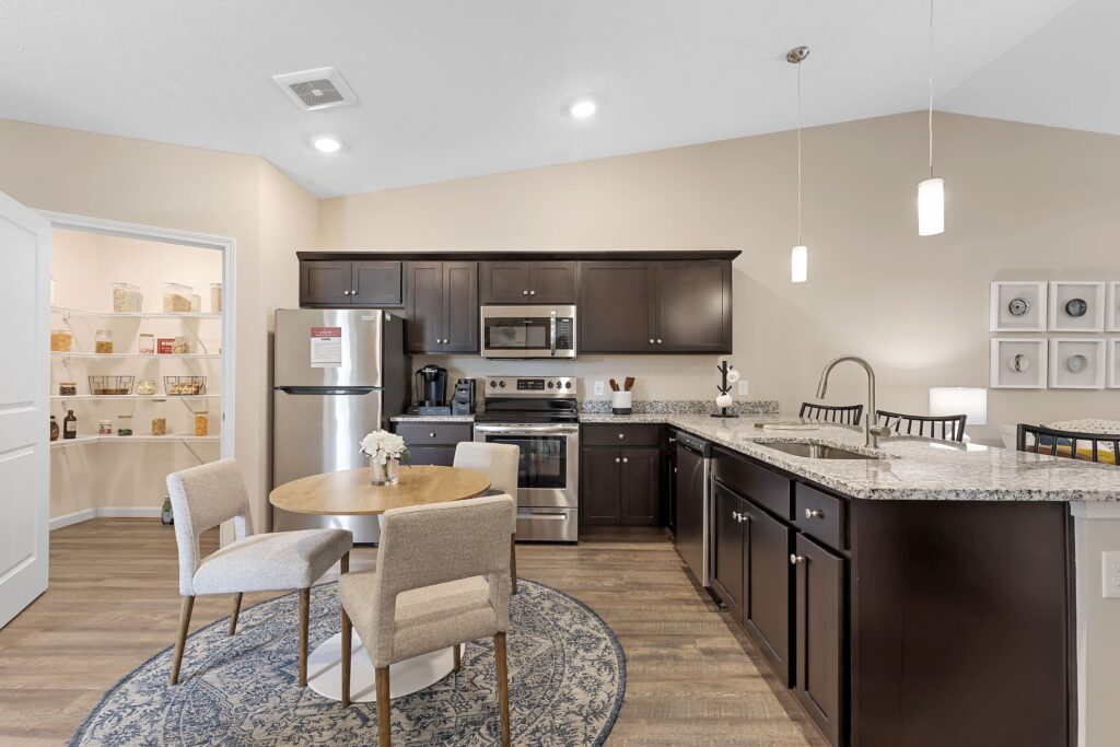 Modern kitchen with dark cabinets, stainless steel appliances, a round dining table with two chairs, granite countertops, pendant lights, and an open pantry in the background.