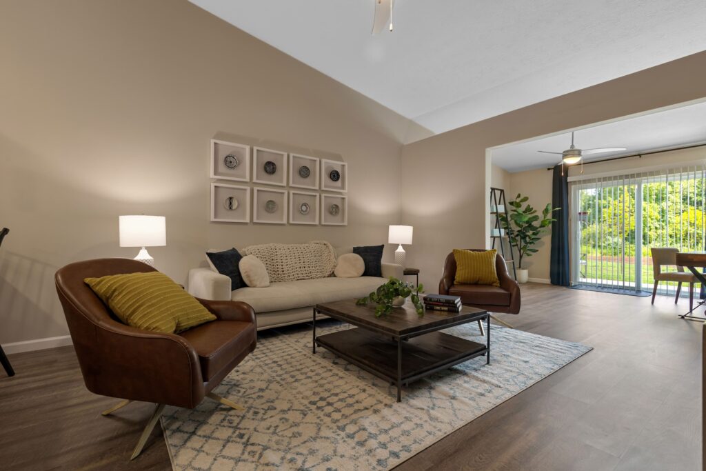 A modern living room with a beige sofa, two brown chairs, a patterned rug, a coffee table, wall art, and large sliding glass doors opening to a green outdoor area.