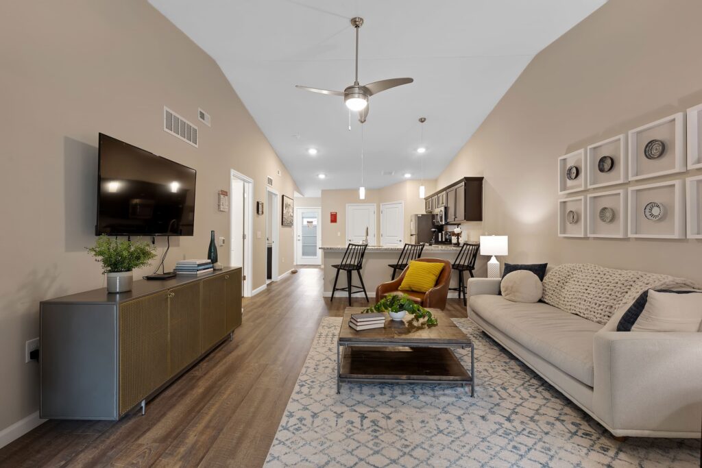Modern living room with beige walls, mounted TV, contemporary furniture, open kitchen, and dining area; décor includes plants, wall art, and patterned rugs.