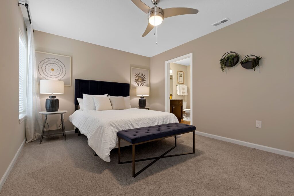 A modern bedroom features a bed with a white comforter, two lamps, a navy bench, beige walls, and décor. An open door leads to a bathroom.
