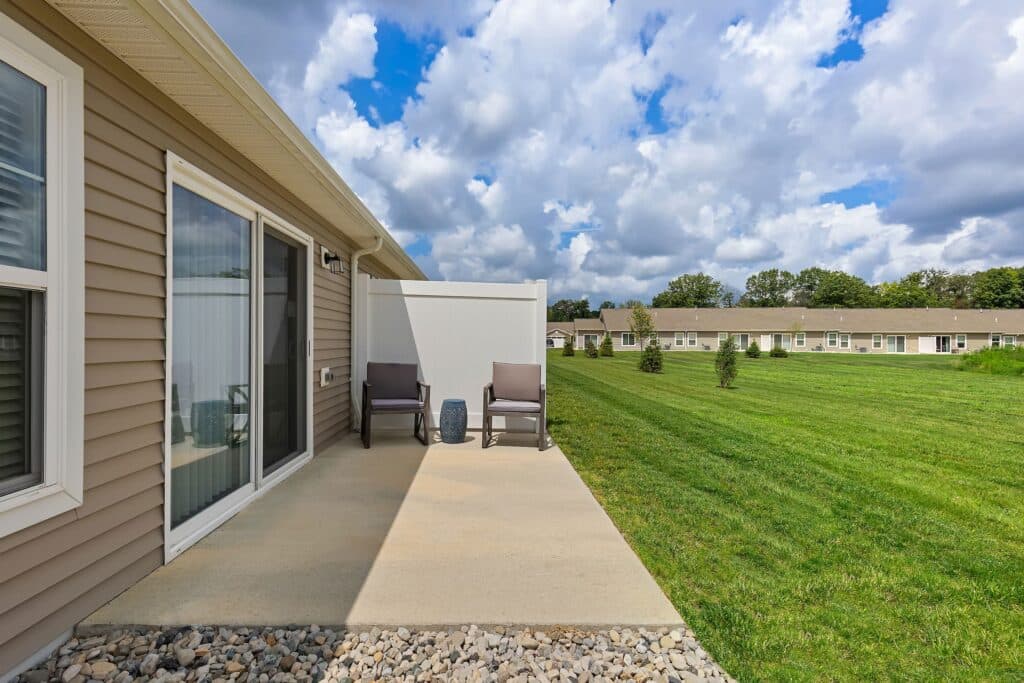 Concrete patio with two chairs and a small table beside a tan house, overlooking a grassy lawn and similar houses in the background under a partly cloudy sky.