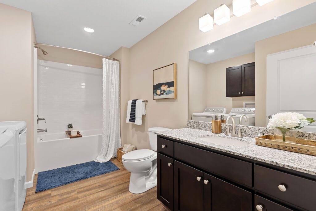 Modern bathroom with a granite countertop, dark wood cabinets, white toilet, shower with white curtain, laundry machines, and wood-look flooring.