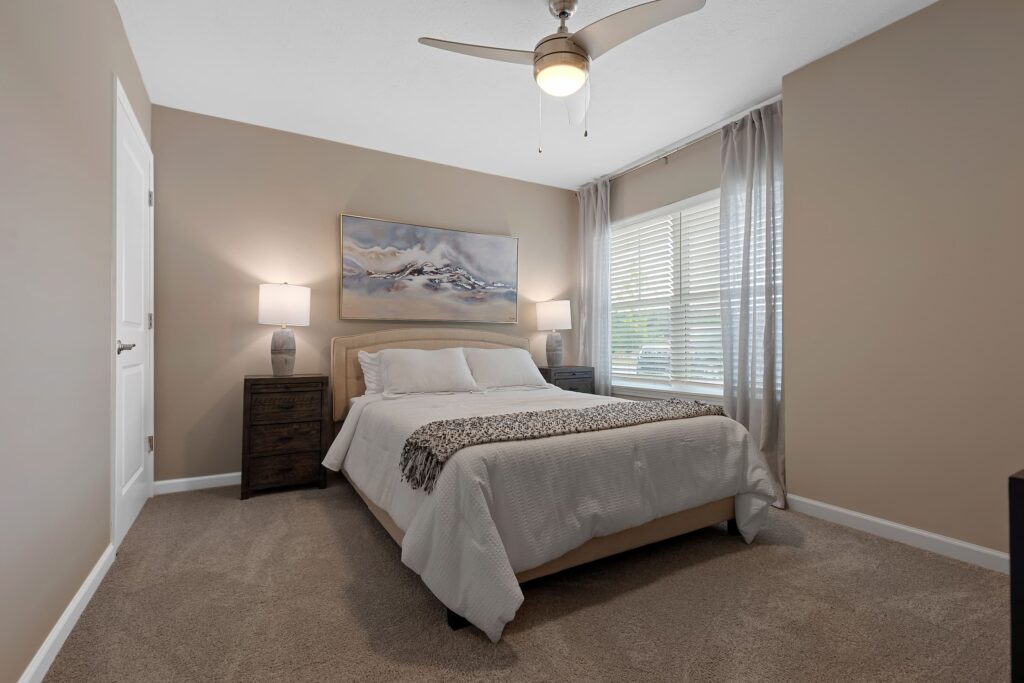 A neatly made bed with white linens is centered between two dark nightstands, each with a lamp. A landscape painting hangs above the bed, and sunlight enters through a window with sheer curtains.