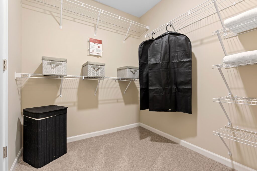 Walk-in closet with wire shelves, three storage bins, a black laundry hamper, a garment bag, and two folded towels on beige carpeted floor.