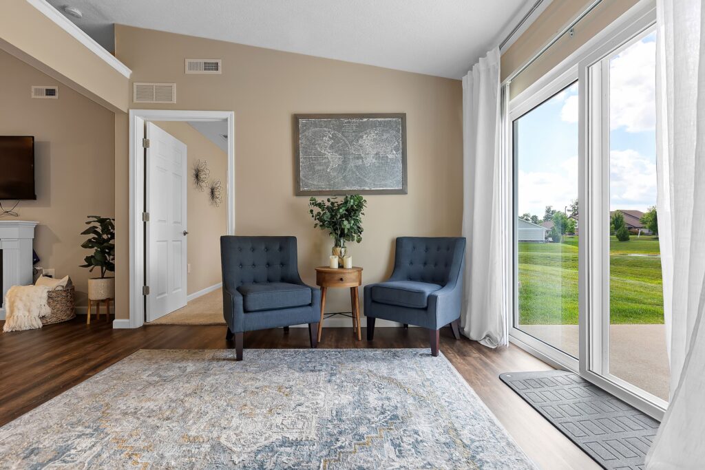 Two blue armchairs and a small wooden table sit near large windows in a bright living room with beige walls and a world map artwork.