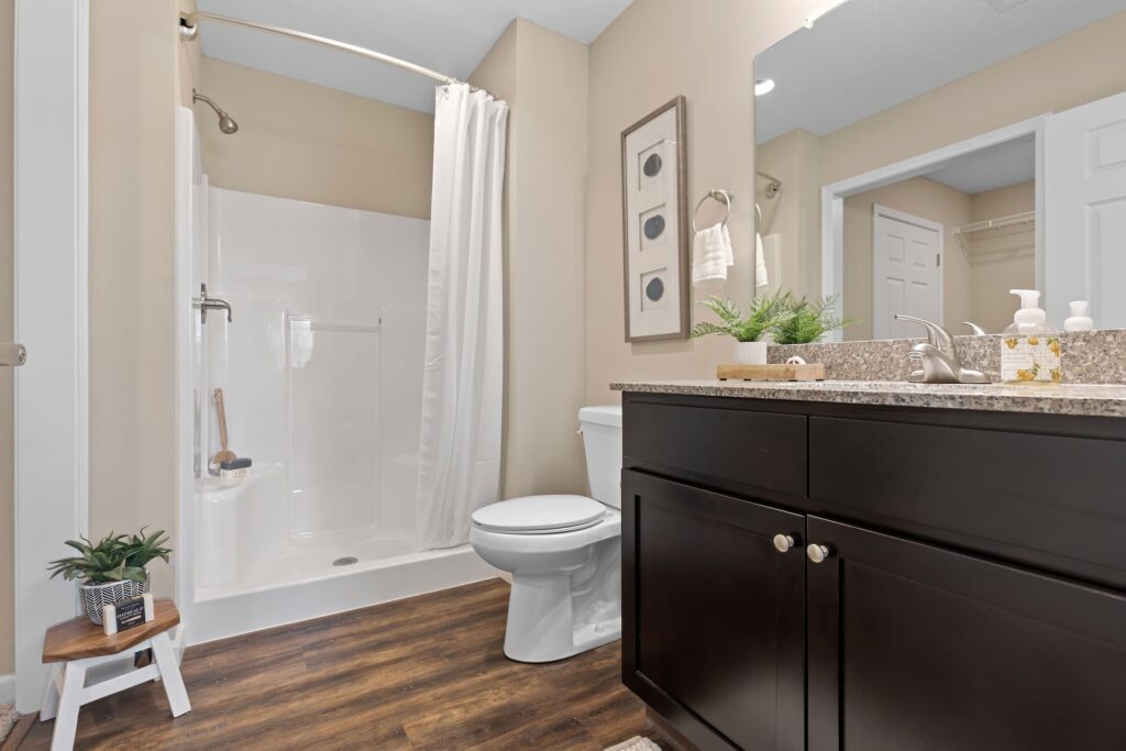 Modern bathroom with a white shower, toilet, dark wood vanity, granite countertop, framed art, and potted plants on a small stool and counter.