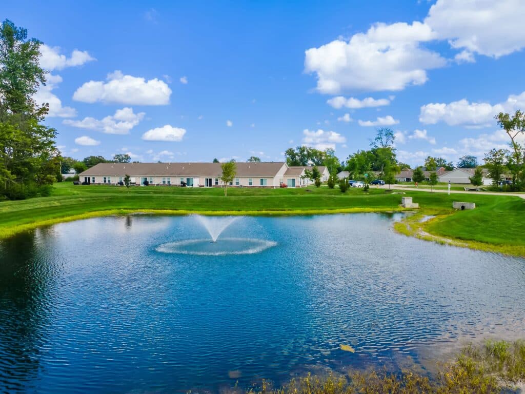 A pond with a central fountain is surrounded by grass, trees, and single-story buildings under a blue sky with scattered clouds.
