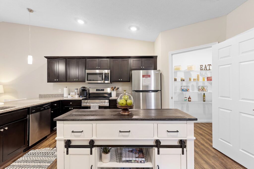 Modern kitchen with dark cabinets, stainless steel appliances, a white island, and an open pantry door revealing shelves with food and decor items.