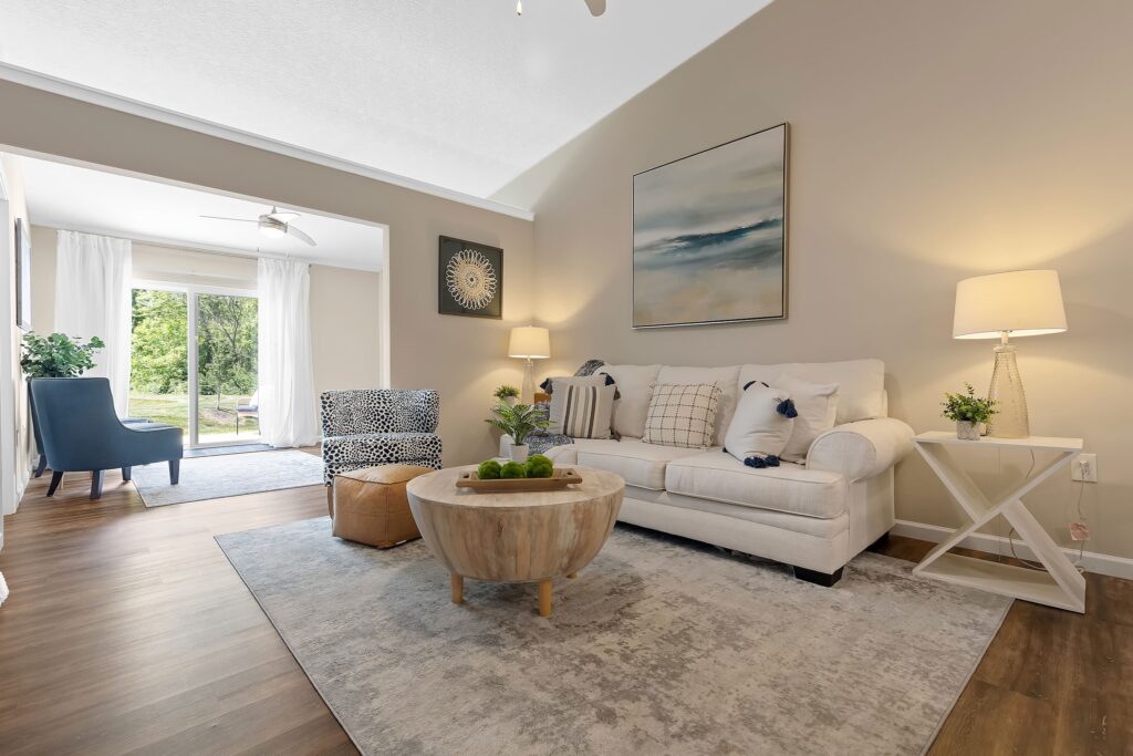 A modern living room with a white sofa, round wooden coffee table, armchair, and lamps, with large windows letting in natural light.