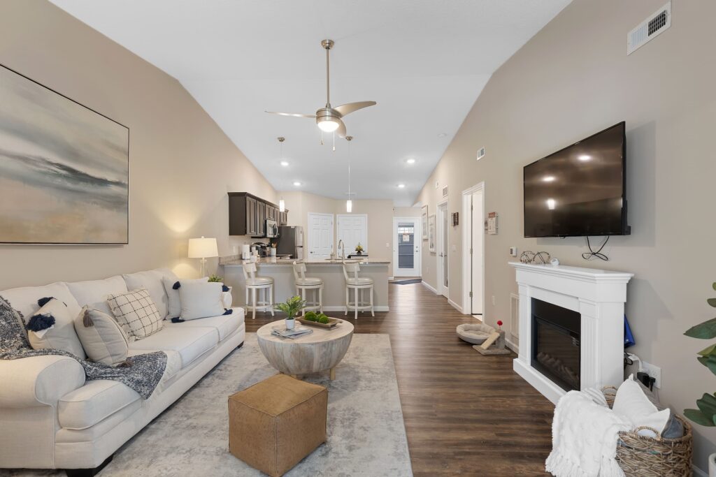 Modern open-concept living room and kitchen with neutral decor, wall-mounted TV above a white fireplace, and a large painting above a light-colored couch.