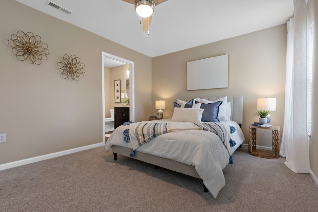 A neatly made bed with blue and white bedding in a carpeted bedroom, featuring beige walls, two lamps, wall art, and a side table with a plant.