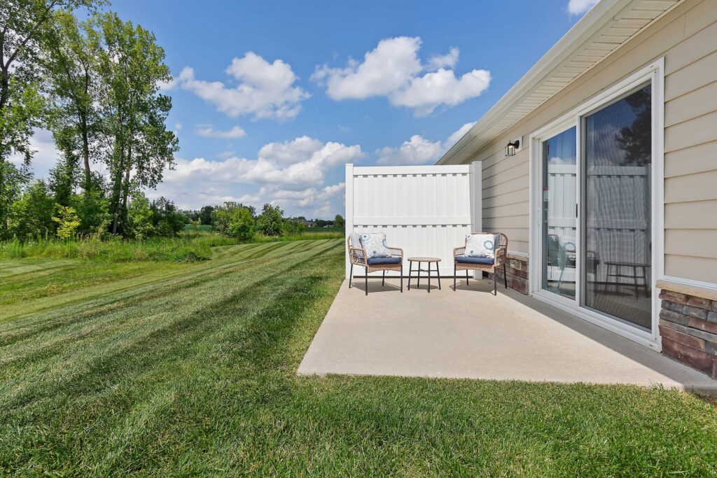 A small concrete patio with two chairs, a side table, and a privacy fence beside a beige house overlooks a grassy lawn and trees under a partly cloudy sky.