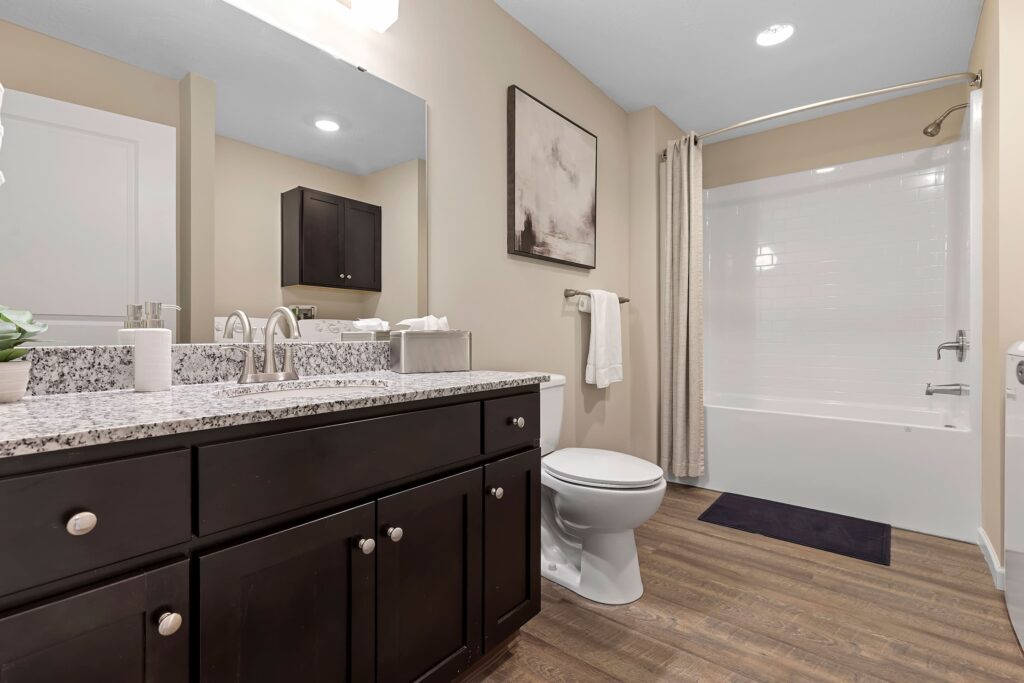 Modern bathroom with dark wood vanity, granite countertop, white sink, toilet, and bathtub with shower curtain; beige walls and wood-look flooring.