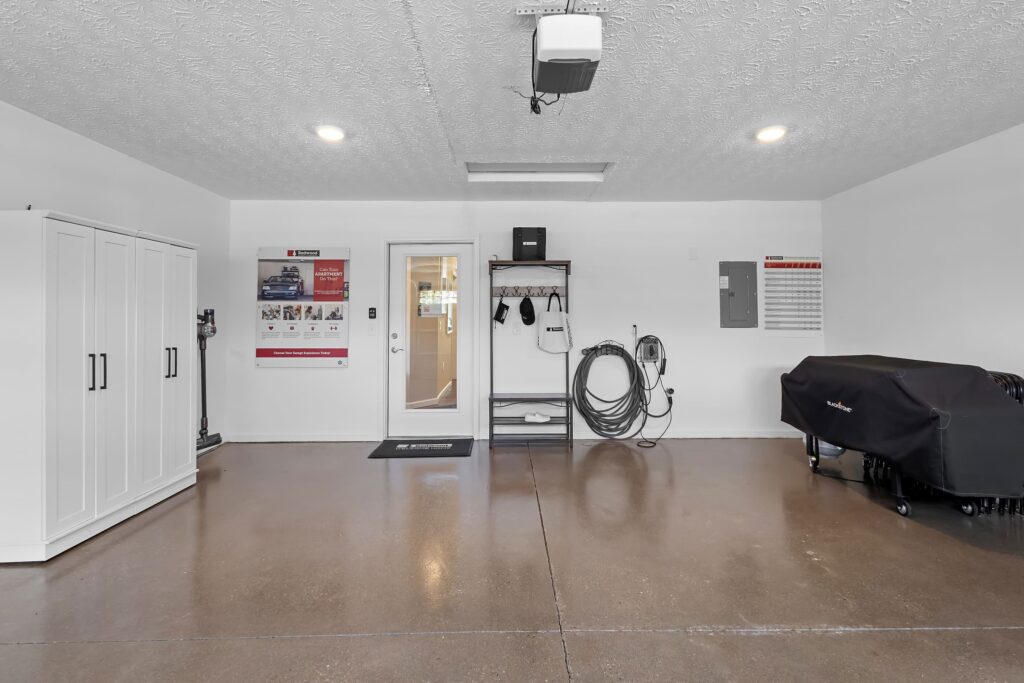 Clean, organized garage with white walls, storage cabinet, utility hooks, hose reel, covered item on right, and posters on wall; door and window in the center background.