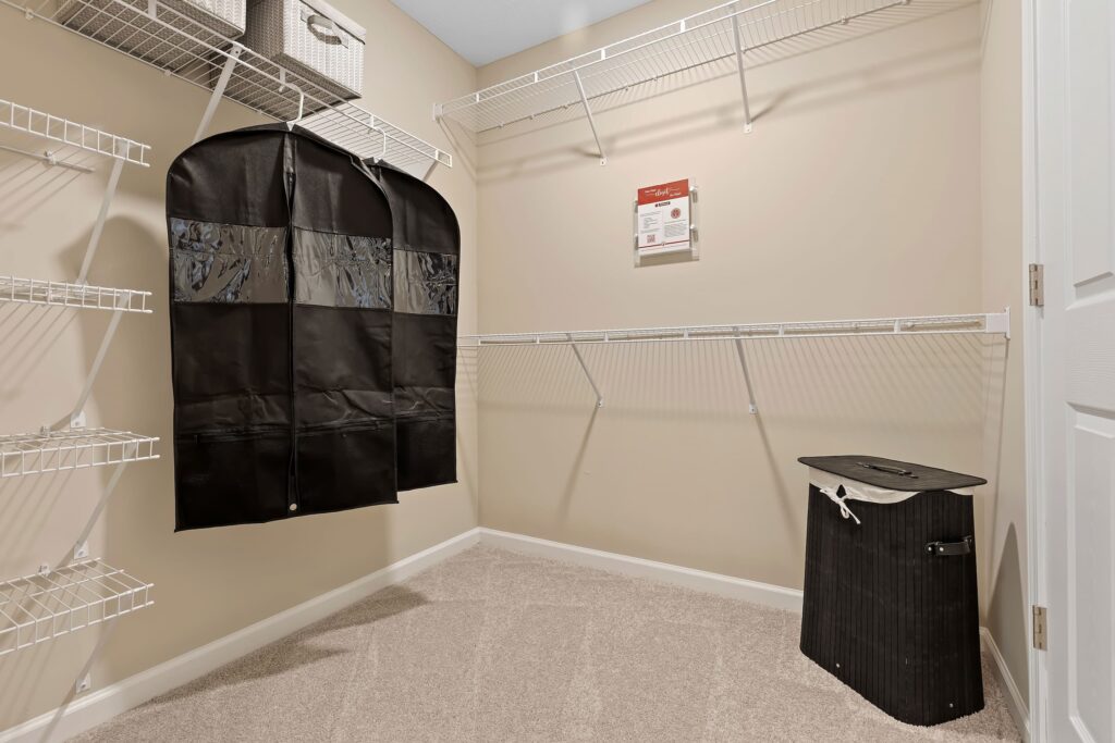 Walk-in closet with beige walls, carpeted floor, wire shelving, a black hanging garment bag, a black laundry hamper, and a small organizer on the upper shelf.