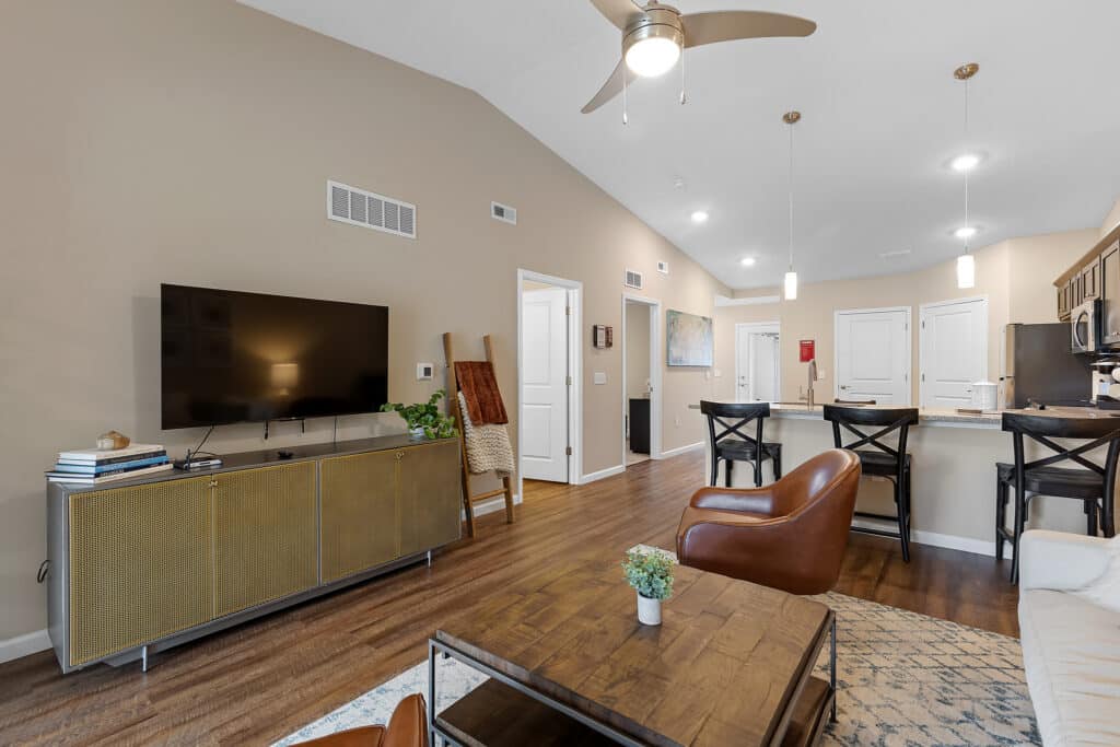 Modern living room with a wall-mounted TV, brown leather chairs, a kitchen island with bar stools, and pendant lighting, featuring neutral tones and wood flooring.