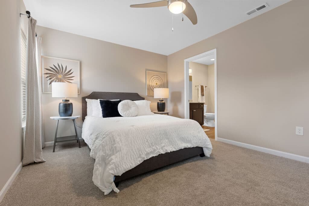 A neatly made bed with white bedding in a neutral-toned bedroom, featuring two bedside tables with lamps and wall art, next to an open door leading to a bathroom.