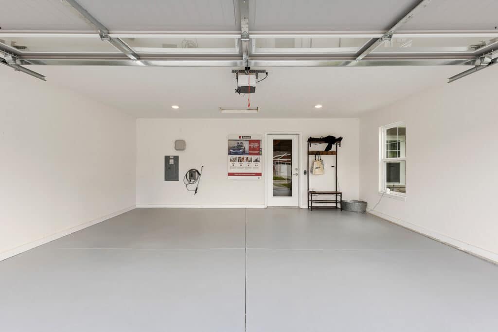 Clean, empty two-car garage with a gray floor, white walls, overhead garage door, shelving unit, utility panel, and two windows. A door leads to an interior room.