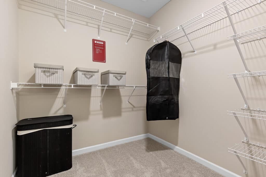 A walk-in closet with wire shelving, three storage bins, a black garment bag, a laundry basket, and a red sign on the beige wall above the shelves.