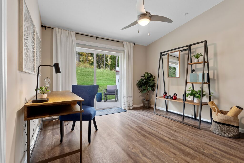 A home office with a wooden desk, blue chair, metal bookshelf, potted plant, and sliding glass doors leading to a patio and grassy yard.
