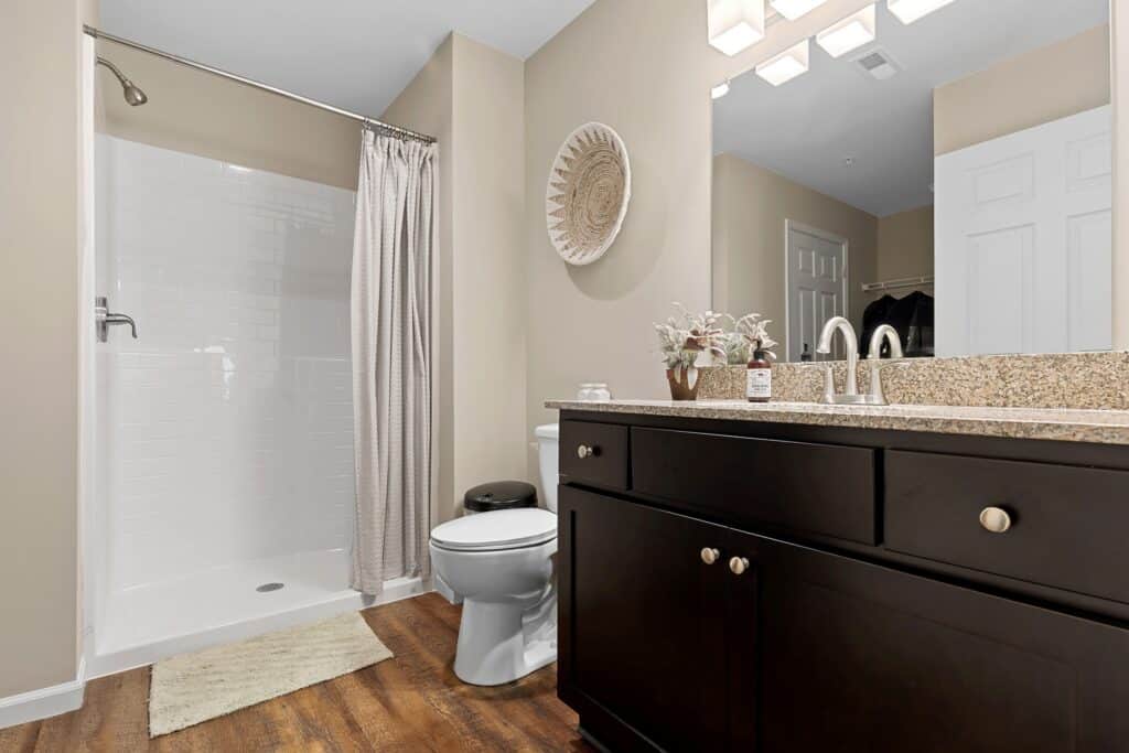 Modern bathroom with a glassless shower, beige walls, dark wood vanity with granite countertop, a toilet, decorative plant, and wall art above the toilet.