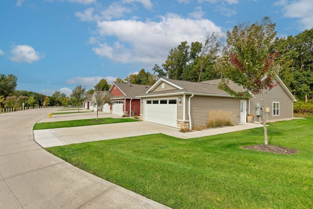 Row of single-story suburban houses with attached garages, well-maintained lawns, and trees lining a quiet residential street under a partly cloudy sky.