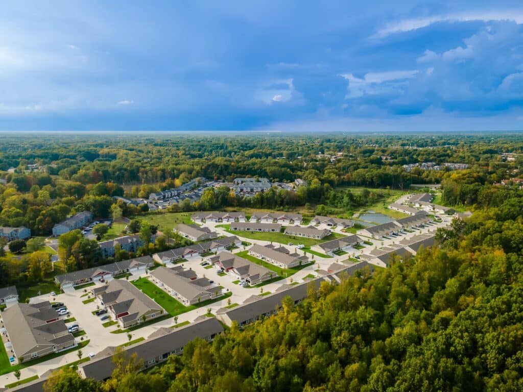 Aerial view of a suburban neighborhood with rows of houses surrounded by dense green trees under a partly cloudy sky.