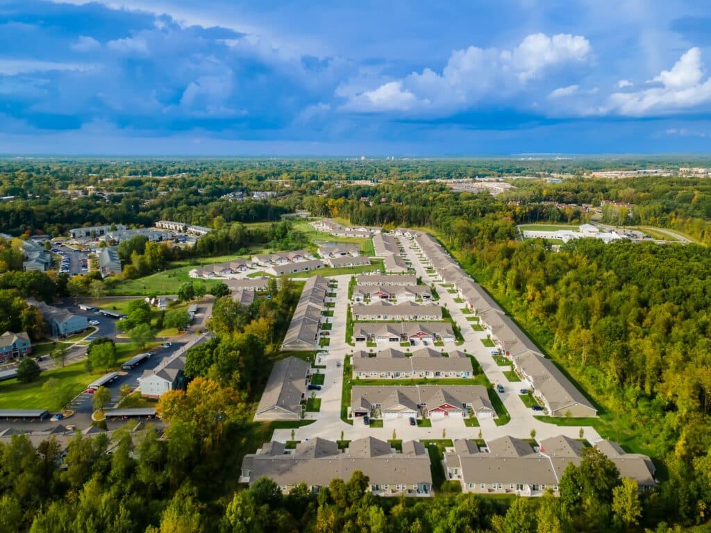 Aerial view of a suburban residential neighborhood with rows of houses, surrounded by trees and greenery under a partly cloudy sky.