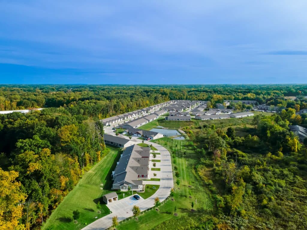 Aerial view of a suburban neighborhood with rows of houses, surrounded by green trees and grassy areas under a blue sky.