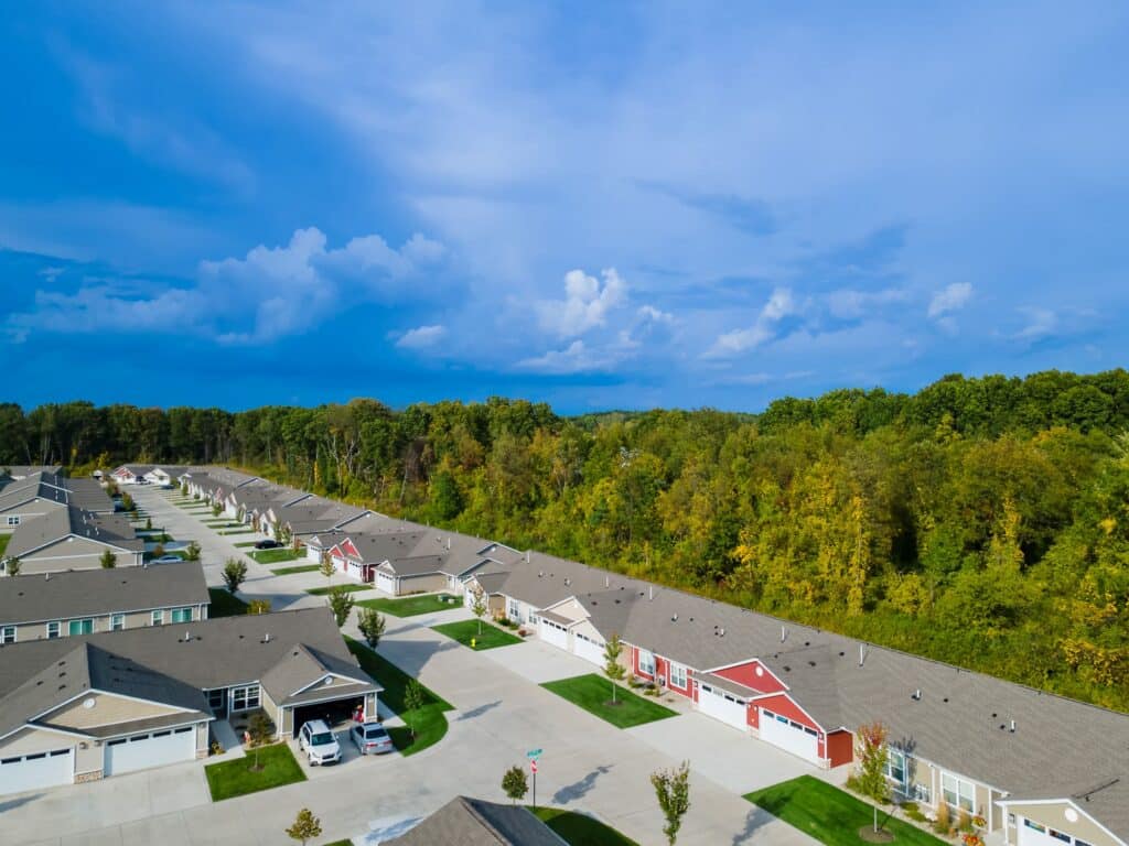 Aerial view of a suburban neighborhood with single-story houses, driveways, parked cars, and a wooded area behind the homes under a partly cloudy sky.
