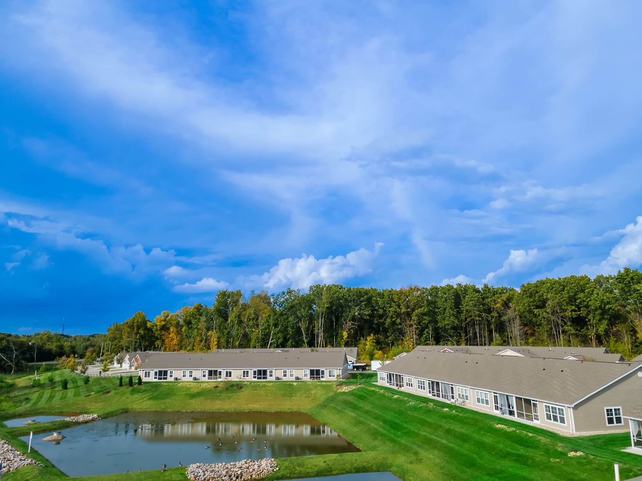 Single-story residential buildings with beige walls and gray roofs sit near a small pond, surrounded by green lawns and trees under a blue sky with scattered clouds.